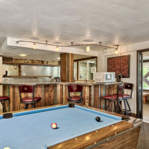 A cozy game room features a pool table, a rustic bar with stools, a microwave, and an exit sign leading to another area.