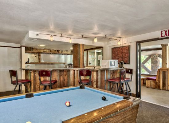 A cozy game room features a pool table, a rustic bar with stools, a microwave, and an exit sign leading to another area.