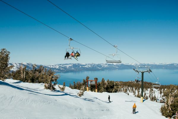 The image shows a ski resort with a chairlift carrying people, skiers on the slopes, snow-covered ground, and a scenic lake in the background.