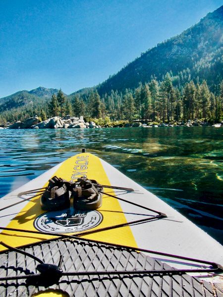 A paddleboard with a pair of shoes is floating on a calm lake, surrounded by forested mountains and clear skies.