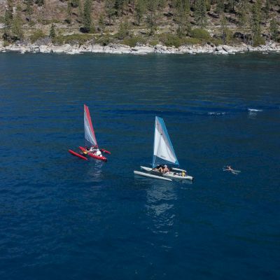 Two sailboats with red and blue sails are sailing on a large body of water, with a swimmer nearby and a rocky shoreline in the background.