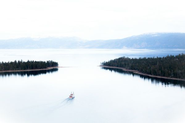 A boat sails between two forested landmasses on a calm lake, surrounded by mountains and an overcast sky in the background.