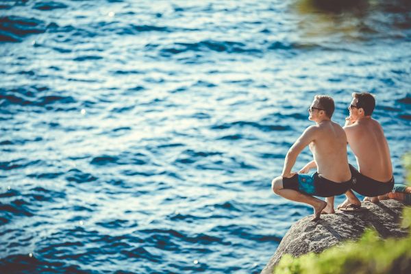 Two shirtless individuals are sitting and squatting on a rock by the water, wearing swim shorts and sunglasses, enjoying the peaceful scenery.