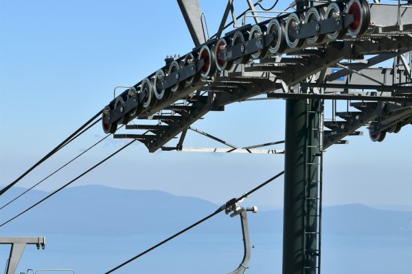 The image shows a gondola lift on a cable, with an ocean or large body of water in the background, and a clear blue sky.