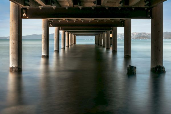The image shows a view from beneath a wooden pier, with columns extending into calm water, mountains visible in the background.