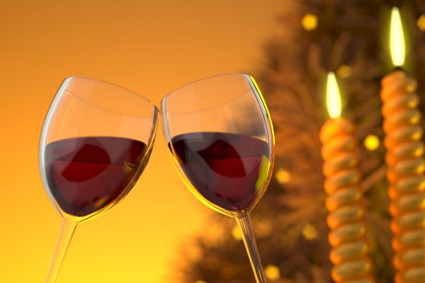 The image shows two wine glasses with red wine clinking together in front of two twisted, lit candles, creating a warm, festive atmosphere.