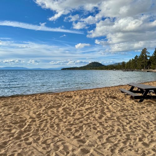 A tranquil sandy beach with a picnic table, calm blue waters, distant hills, and a sky dotted with clouds. Trees line the shore in the background.