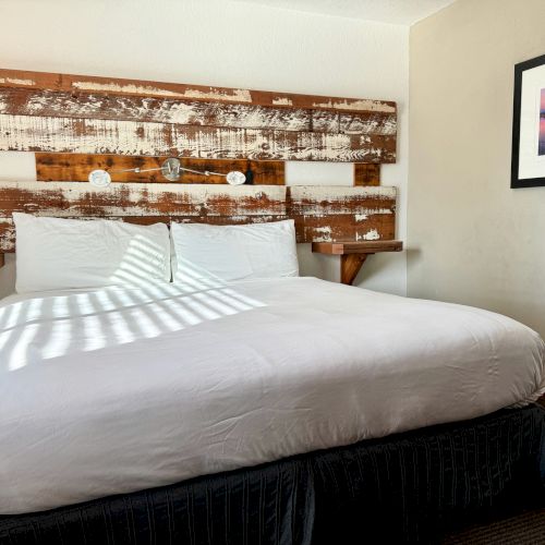 A hotel room with a king-sized bed, rustic wooden headboard, wall sconces, a framed picture, and sunlight streaming in through a window.