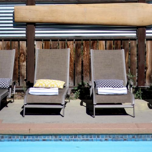 Four lounge chairs with cushions, arranged by a swimming pool near a wooden fence with a surfboard mounted above the chairs.