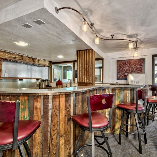 The image shows a rustic bar area with wooden countertops and red cushioned bar stools under industrial-style lighting, featuring a cozy seating area.